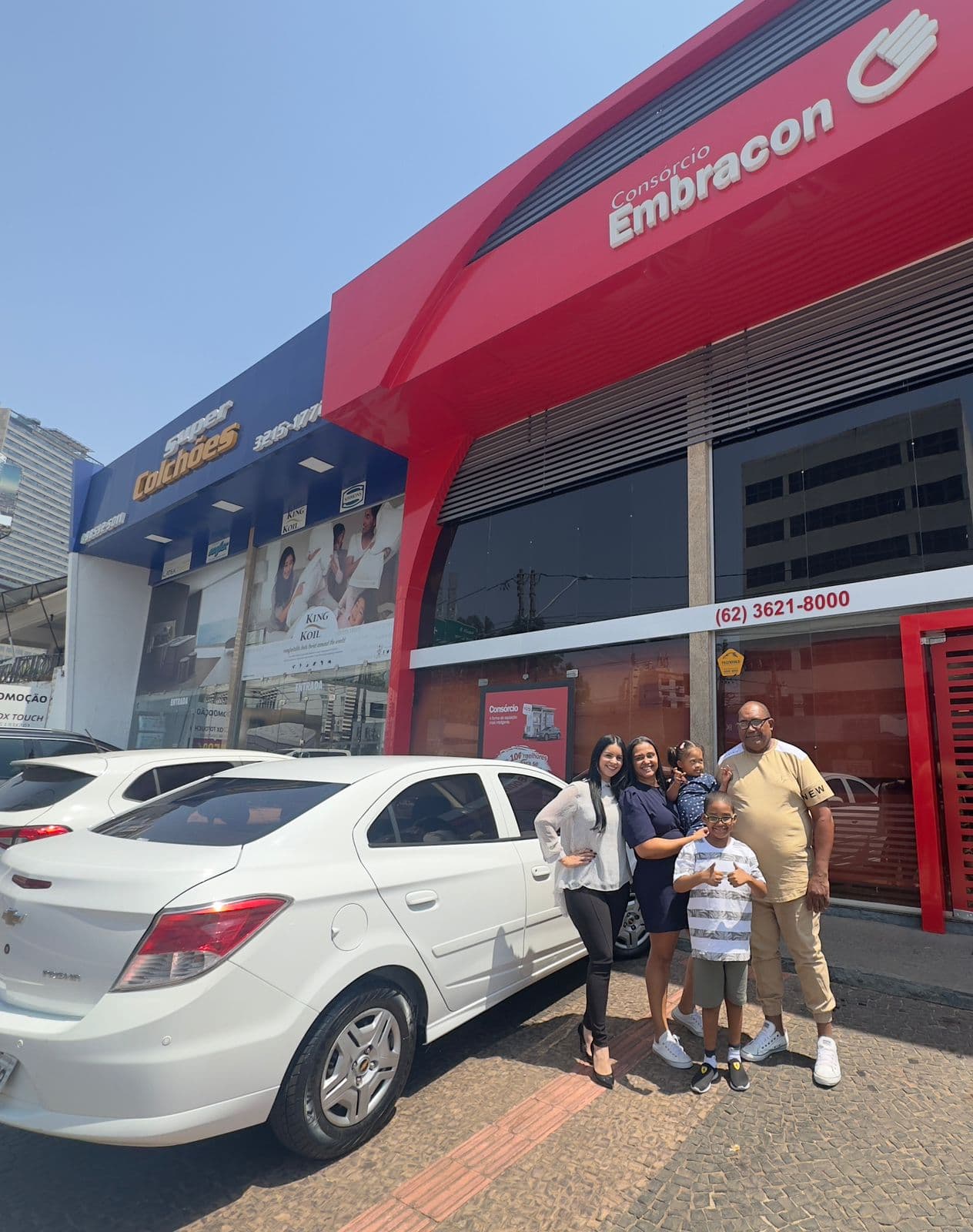 Faça como o Everaldo e garanta uma vida melhor para a sua família com o Consórcio Embracon! Everaldo cliente do consórcio Embracon e sua família em frente a uma agência Embracon